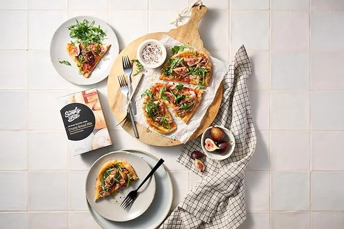 Two slices of pizza on plates with a cutting board featuring a whole pizza, figs, and a napkin on a tiled surface.
